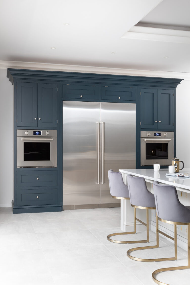Hague Blue cabinetry frames a large Sub-Zero fridge and twin Wolf ovens, with grey velvet stools at the island