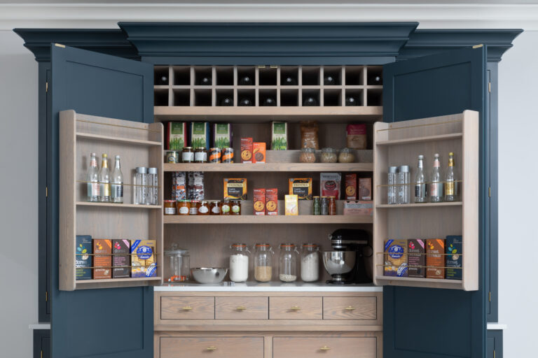 Open Hague Blue pantry cupboard with timber shelving, wine racks, drawers and brass handles, fully stocked with jars and groceries