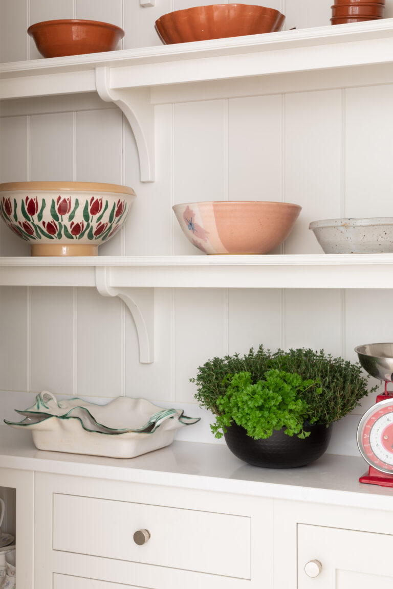 Open shelving above a panelled dresser displays ceramic bowls, a dish, and a potted herb plant