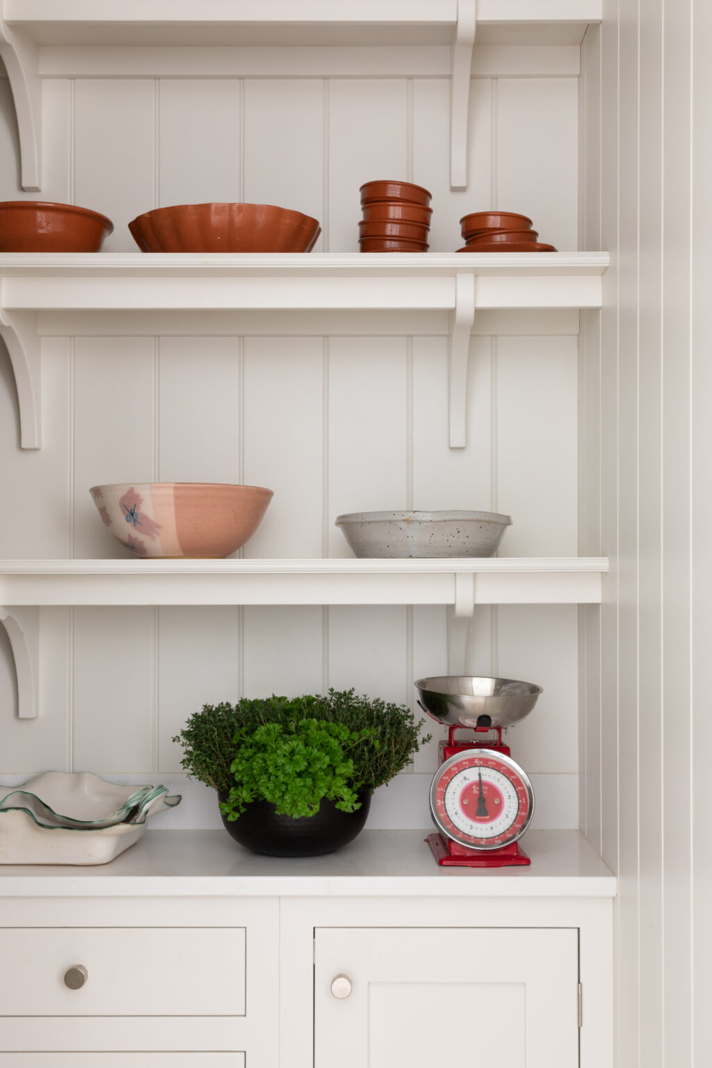 Open painted kitchen shelves display terracotta bowls, ceramic dishes, a potted herb and a vintage red kitchen scale