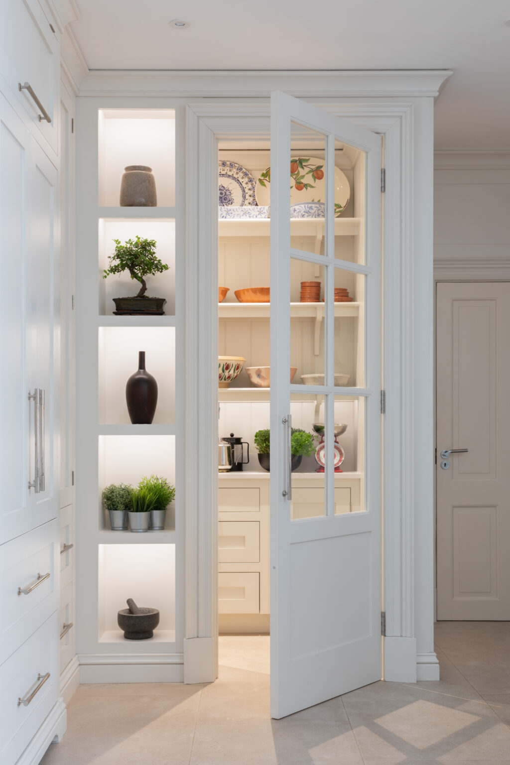 Glazed pantry door opens onto shelved storage with crockery and bowls, beside lit display niches