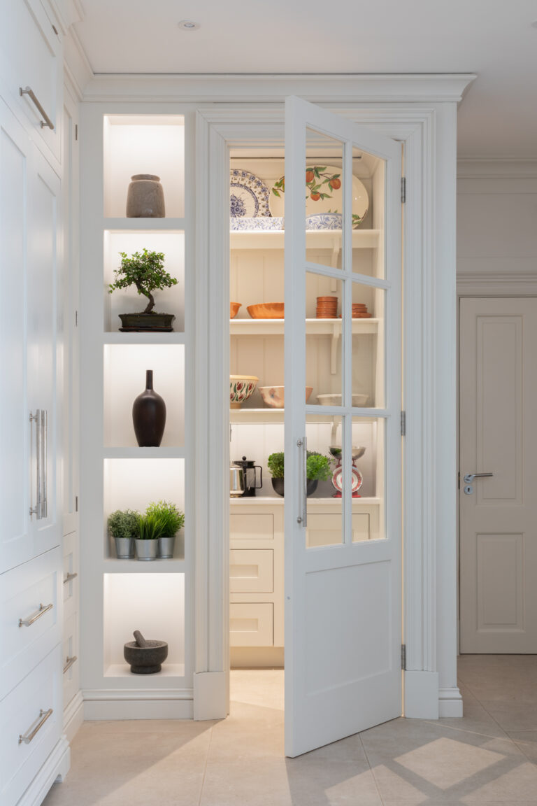 Glazed pantry door opens onto shelved storage with crockery and bowls, beside lit display niches