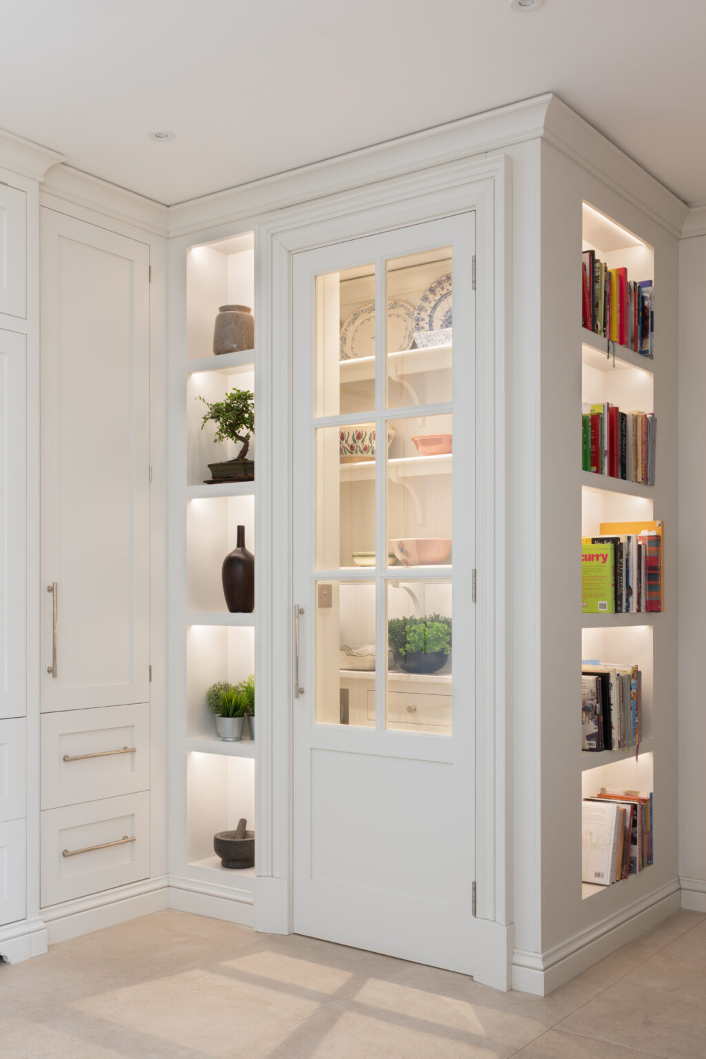 Glazed pantry door with illuminated open shelving either side, displaying crockery, cookbooks and decorative ornaments