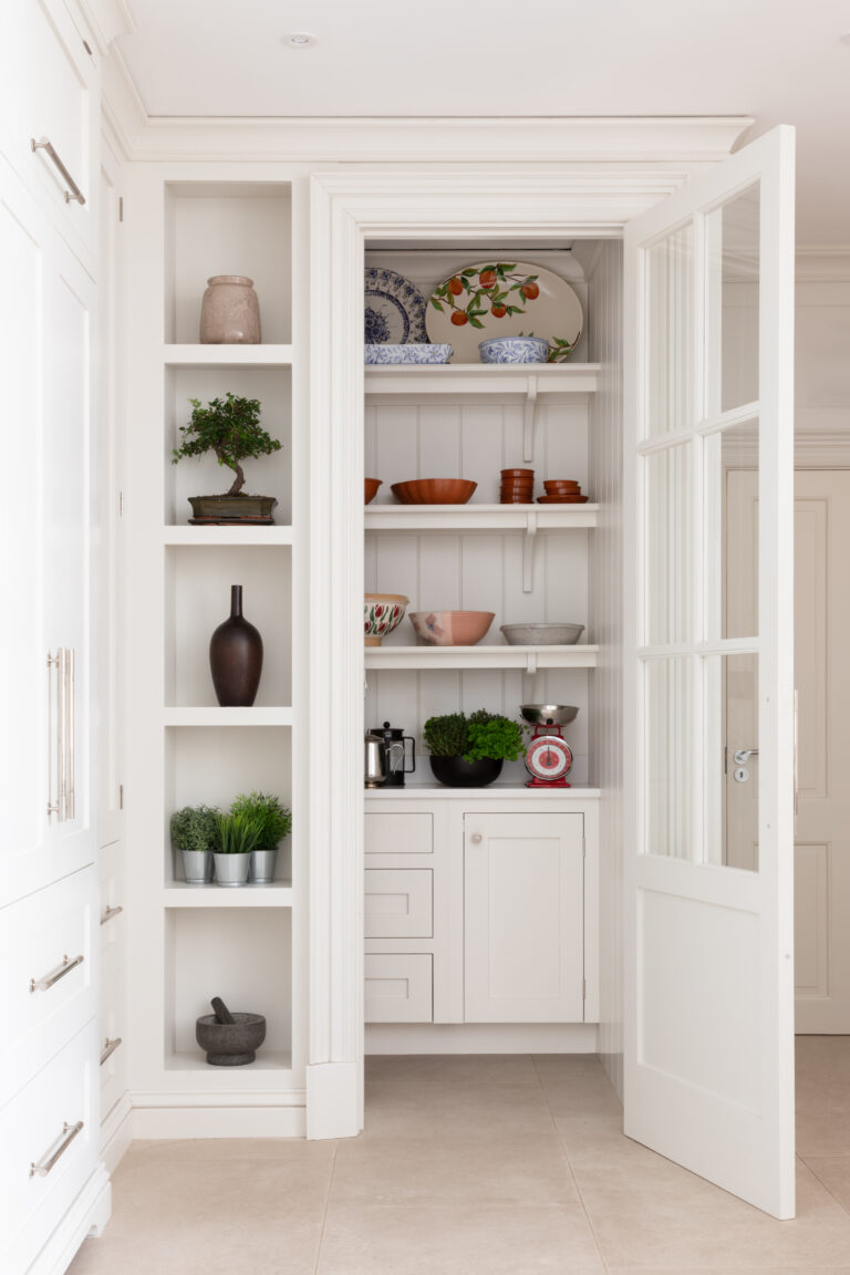 Glazed door opens to a bespoke walk-in pantry with open shelving displaying crockery, bowls and plants