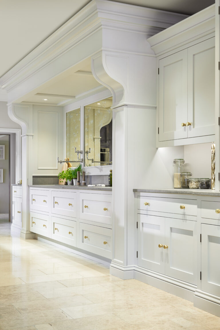 Bespoke painted cabinetry with brass handles, panelled drawers and ornate cornicing frames a mirrored splashback and worktop