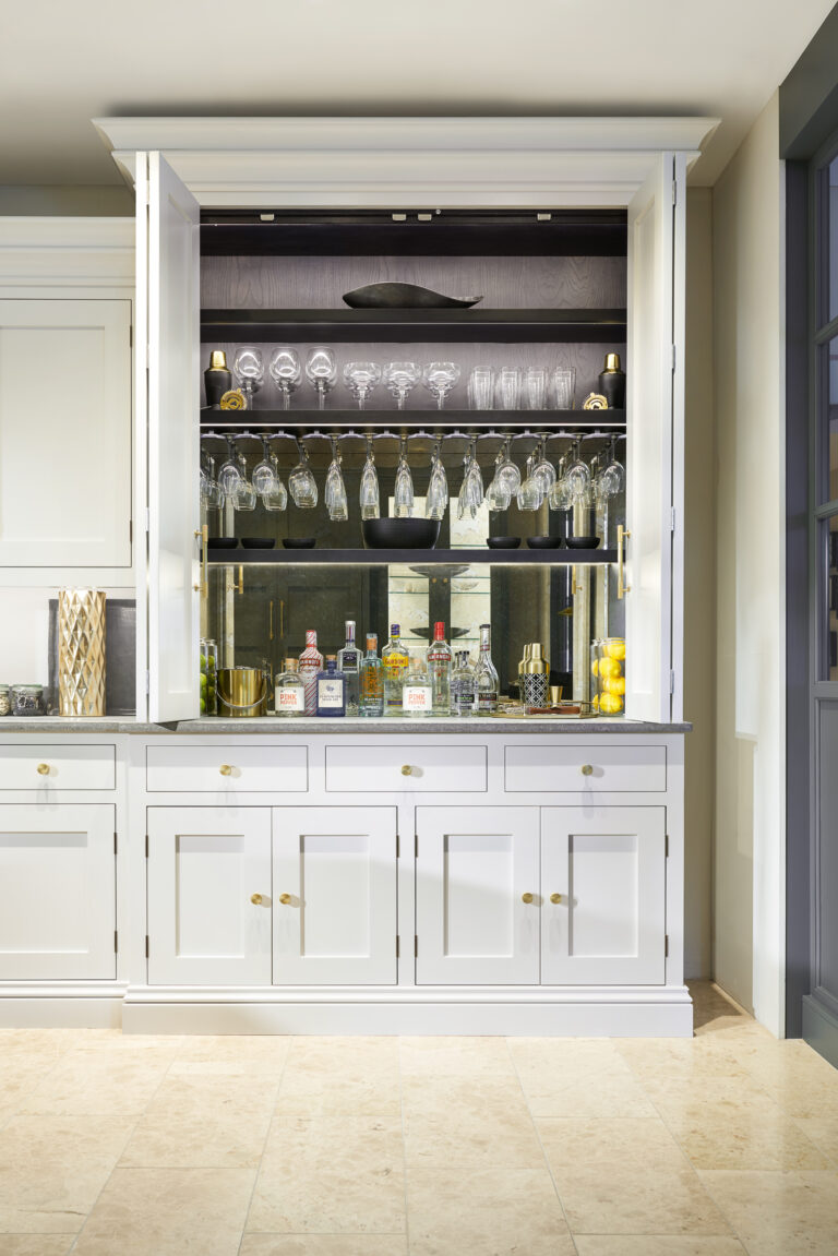 Open wet bar cabinet with mirrored back, glassware, hanging glasses and brass handles above cupboards and drawers