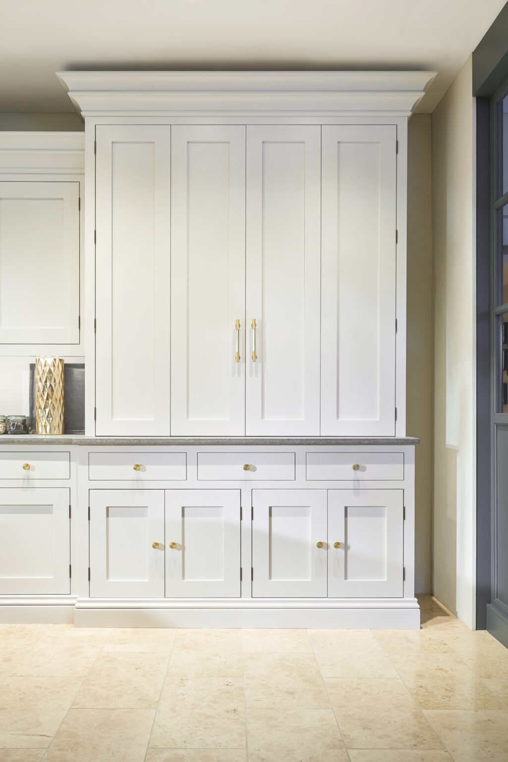 Tall painted pantry cupboard with panelled doors, drawers and polished brass handles, set against a pale wall