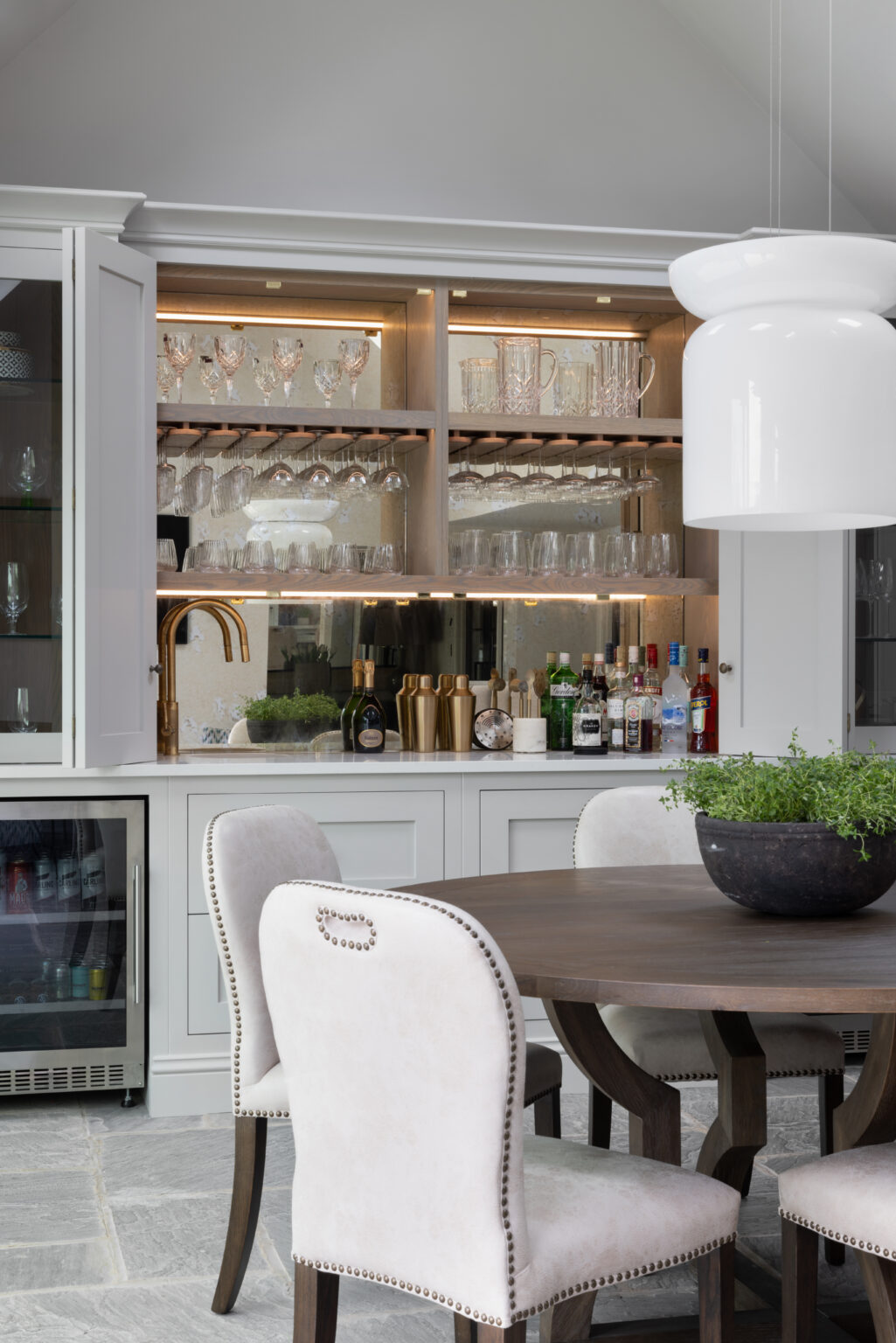 Bespoke home bar with glassware shelving and brass tap, beside a dining table with upholstered chairs