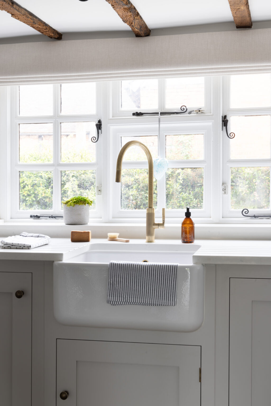 Butler sink with brass tap beneath a timber-beamed window, set into pale painted cabinetry