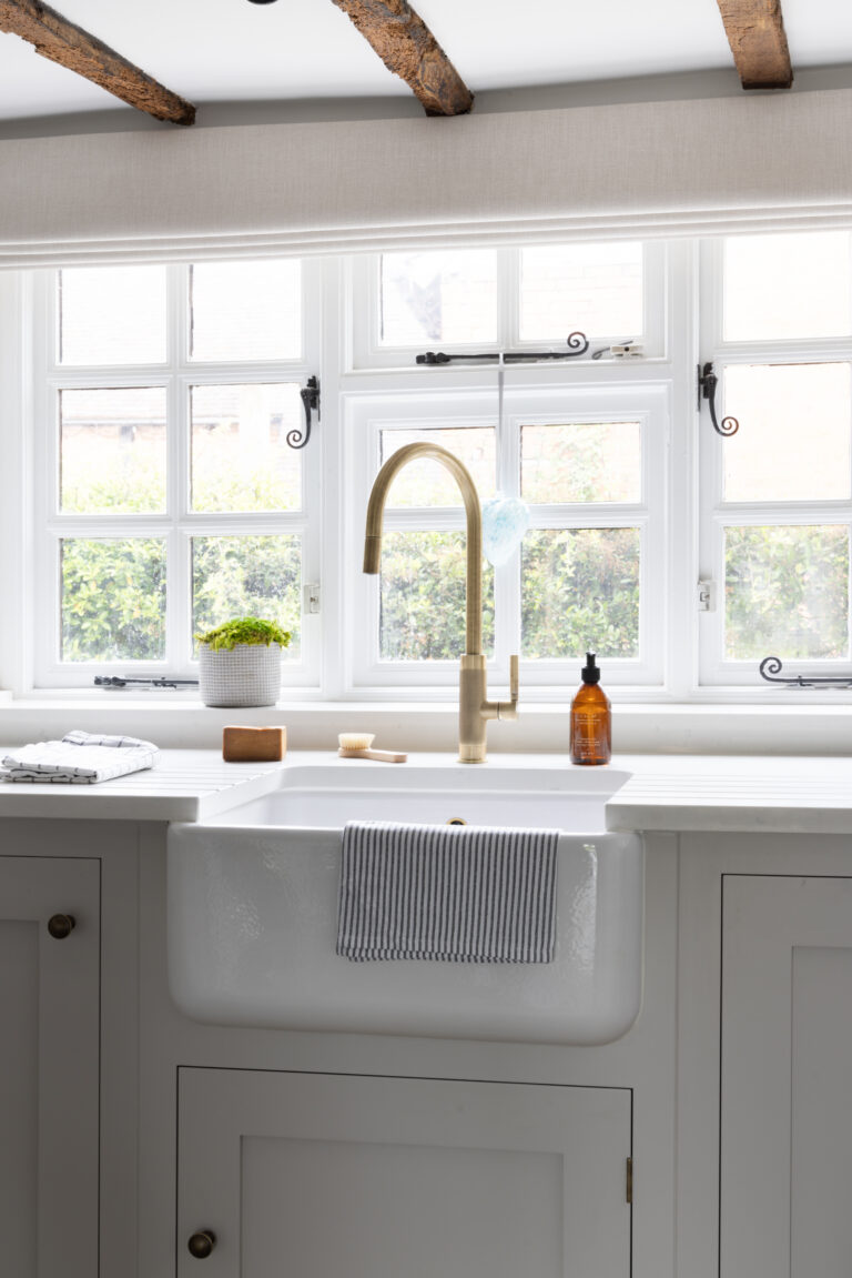 Butler sink with brass tap beneath a timber-beamed window, set into pale painted cabinetry