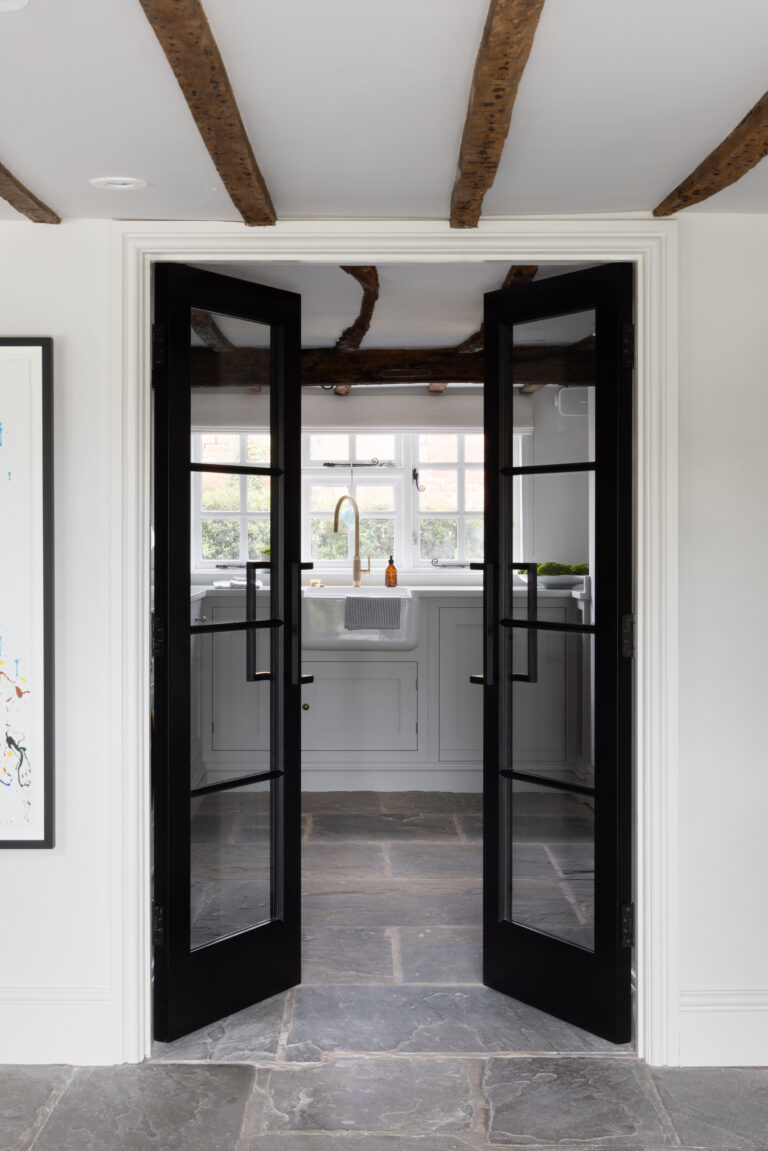 Black-framed glazed double doors open to a kitchen with a Belfast sink, exposed beams and flagstone flooring