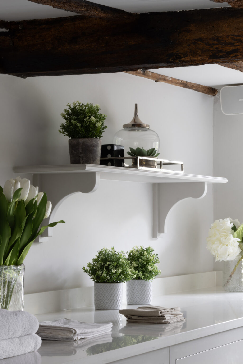 White bracket shelf beneath exposed timber beams, styled with plants, jars and folded linens on the worktop