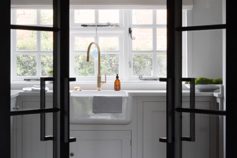 Belfast sink with brass tap beneath leaded windows, seen through black-framed glazed doors into the utility area