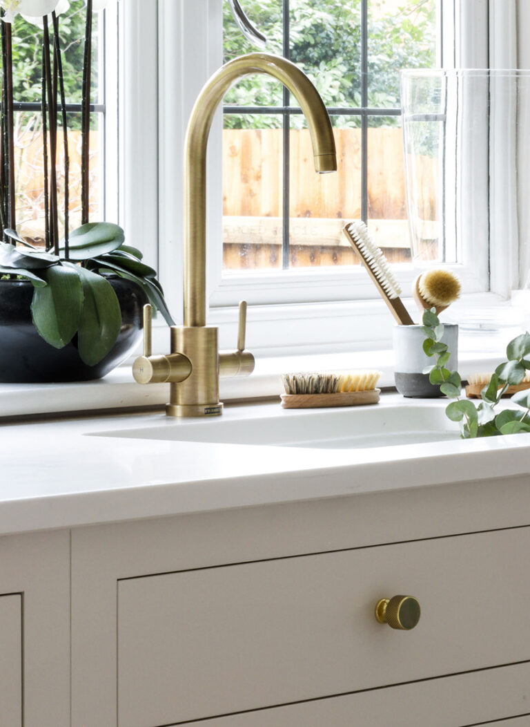 Brass mixer tap and knurled handle above a white sink, set into a pale shaker-style cabinet by the window