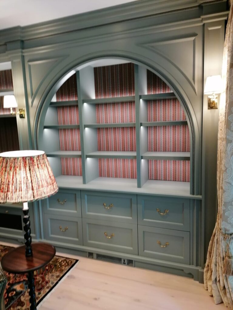 Living room alcove with a built-in arched bookcase and lower drawers, brass handles, and striped fabric-lined shelving
