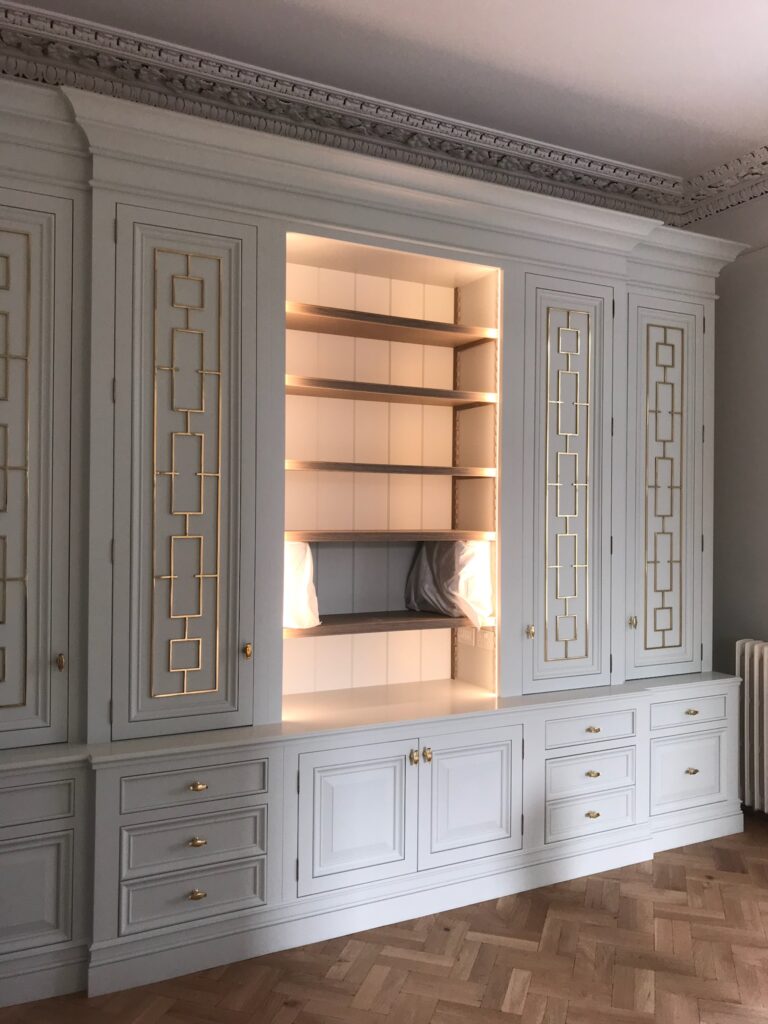 Built-in bespoke wall unit with glazed brass fretwork doors, drawers, cupboards and an illuminated open shelving display section