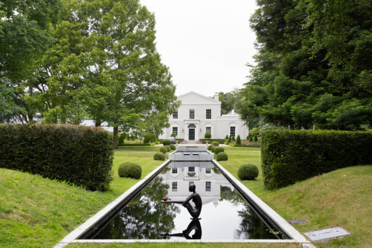 Grand white stately home viewed across a long reflecting pool with a bronze sculpture and manicured hedges