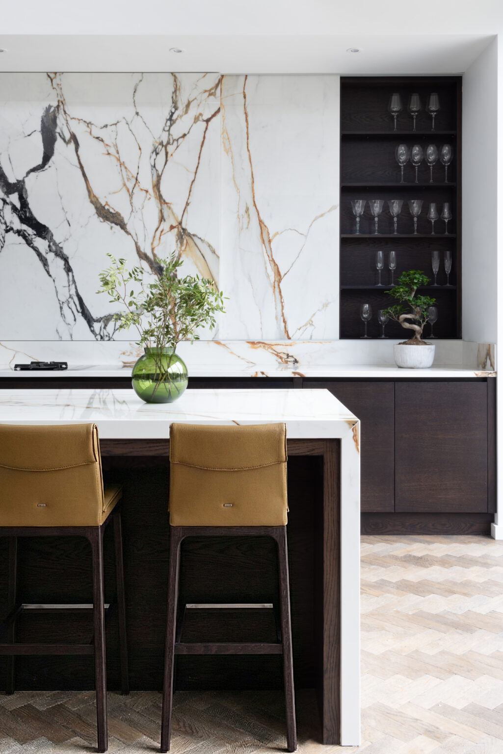 Kitchen island with mustard bar stools, a moving stone wall, and open shelving displaying glassware