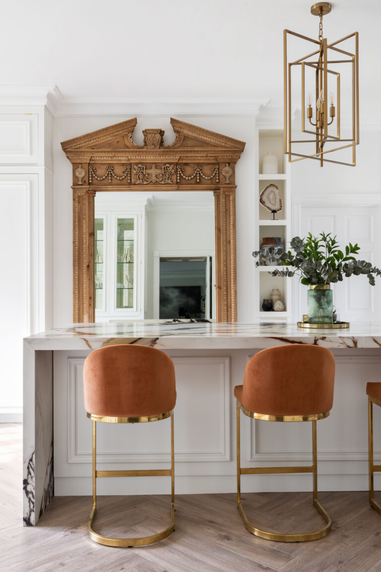 Antique carved mirror above an XTONE island with burnt-orange velvet stools and brass legs