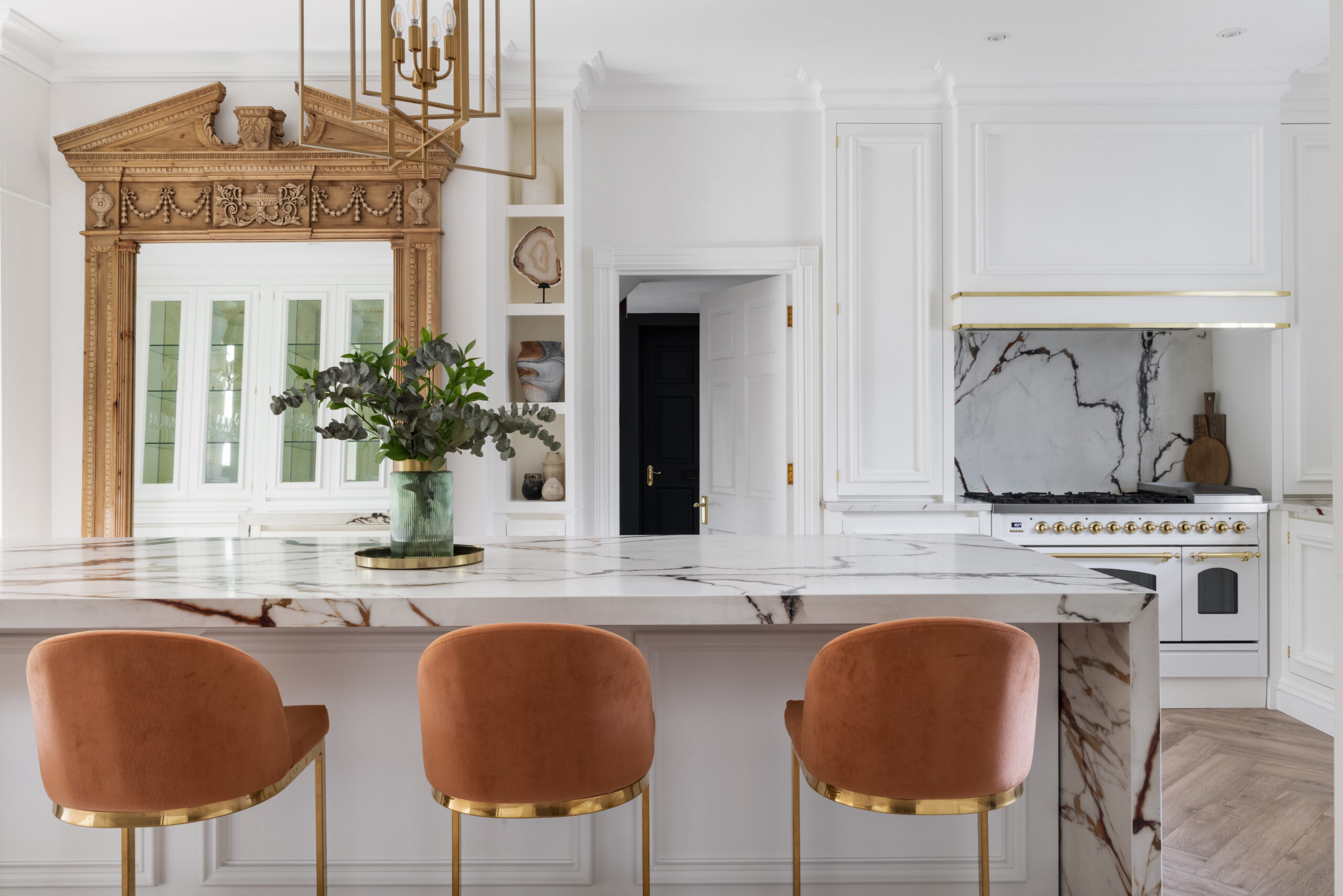 Antique mirror and XTONE island with burnt-orange velvet stools and a professional-grade cooking range beyond