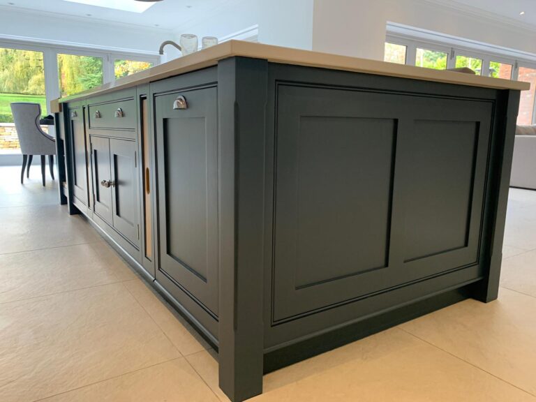 Large kitchen island with panelled cabinet doors, brass handles and a pale worktop, set in a bright open room