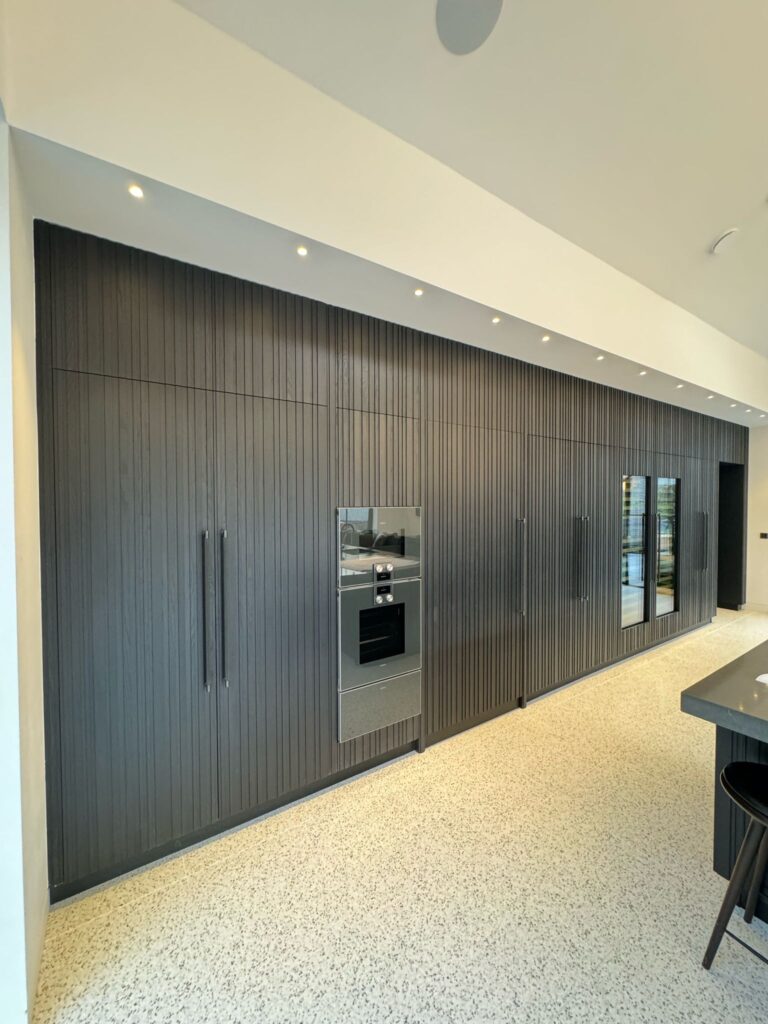 Kitchen wall of full-height ribbed panelled cabinetry with integrated oven, long handles and recessed ceiling spotlights above