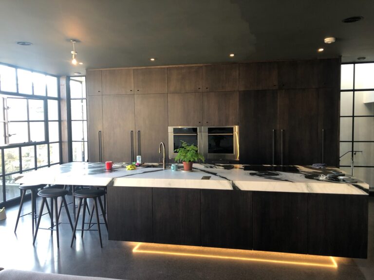 Large kitchen island with veined stone worktop, dark cabinetry, bar stools and built-in ovens along the wall