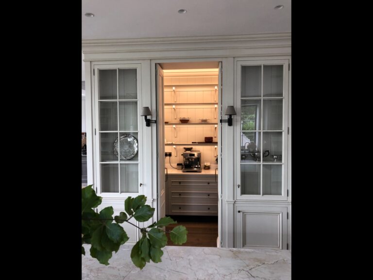 Glazed display cabinets flank an open door revealing a lit pantry with shelving, drawers and a coffee machine