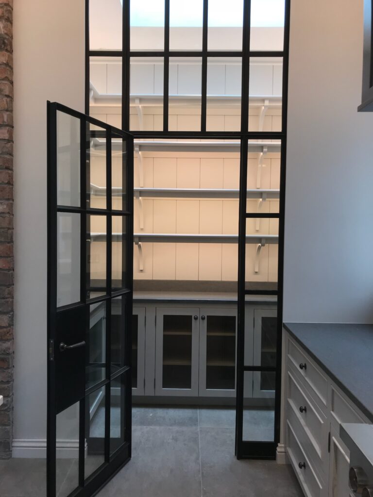Crittall-style glazed door opens onto a pantry with tall shelving, glass-fronted cabinets and a stone worktop
