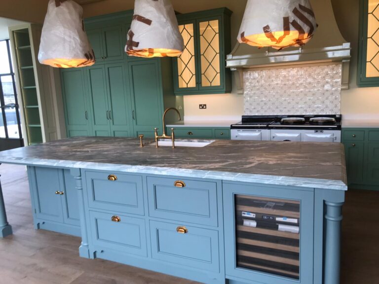 Kitchen island with stone worktop, brass tap and fittings, green cabinetry and range cooker beyond, pendant lights still wrapped