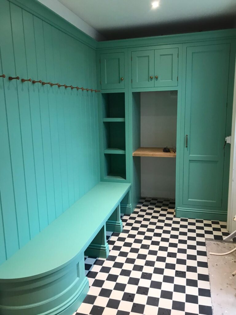 Boot room with green panelling, fitted bench, coat pegs and tall cupboards above a chequered black and white floor