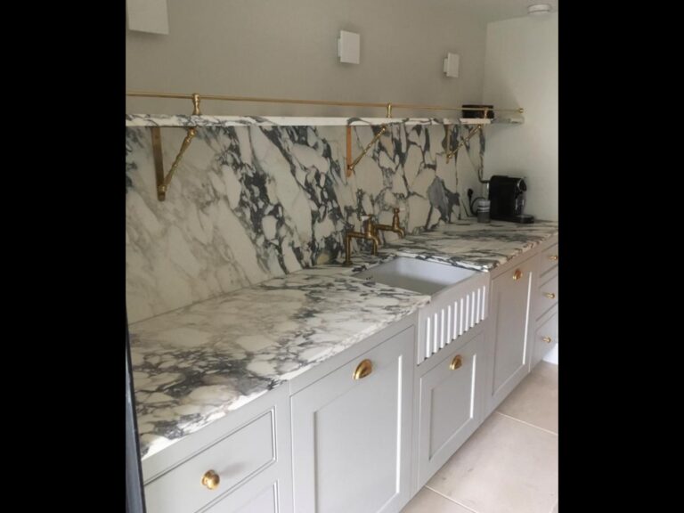 Kitchen run with stone worktop and splashback, white cabinetry, brass handles, taps and shelf brackets, and a butler sink