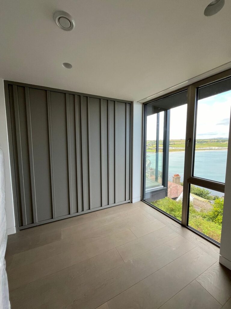 Bedroom with fitted fluted wardrobe doors beside a full-height window overlooking the coastline and harbour