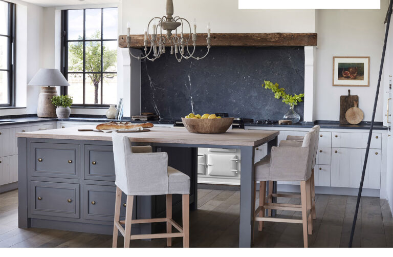 Kitchen island with grey cabinetry and timber worktop, upholstered bar stools, and a dark stone splashback behind the range