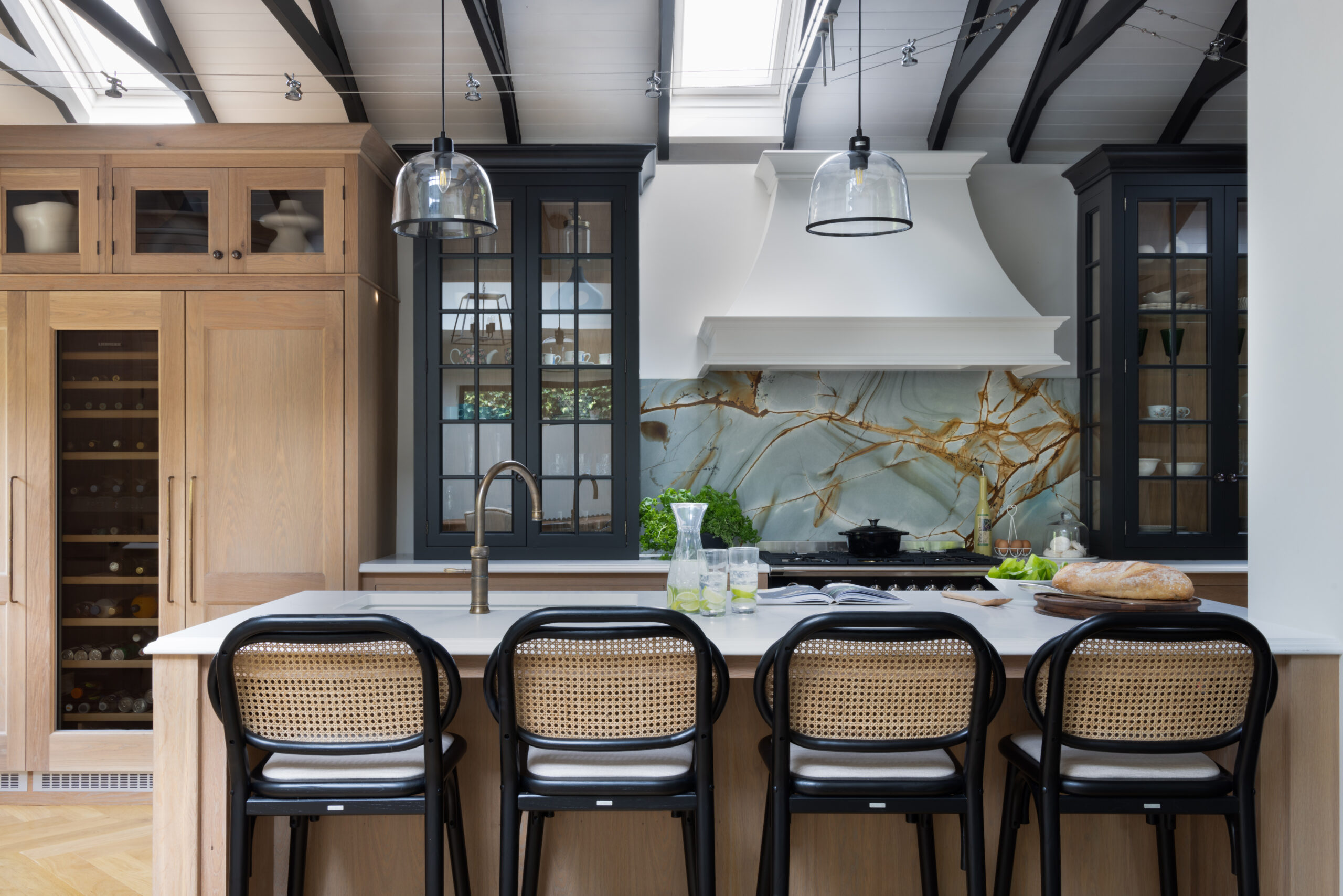 Kitchen island with cane-back stools beneath vaulted beams, facing a backlit stone splashback and white canopy