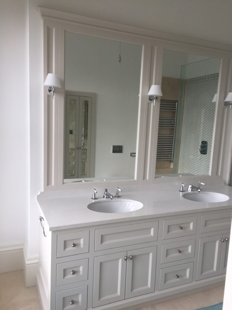 Bathroom vanity unit with twin inset basins, tall mirrors, wall lights and panelled cabinet doors beneath a stone worktop
