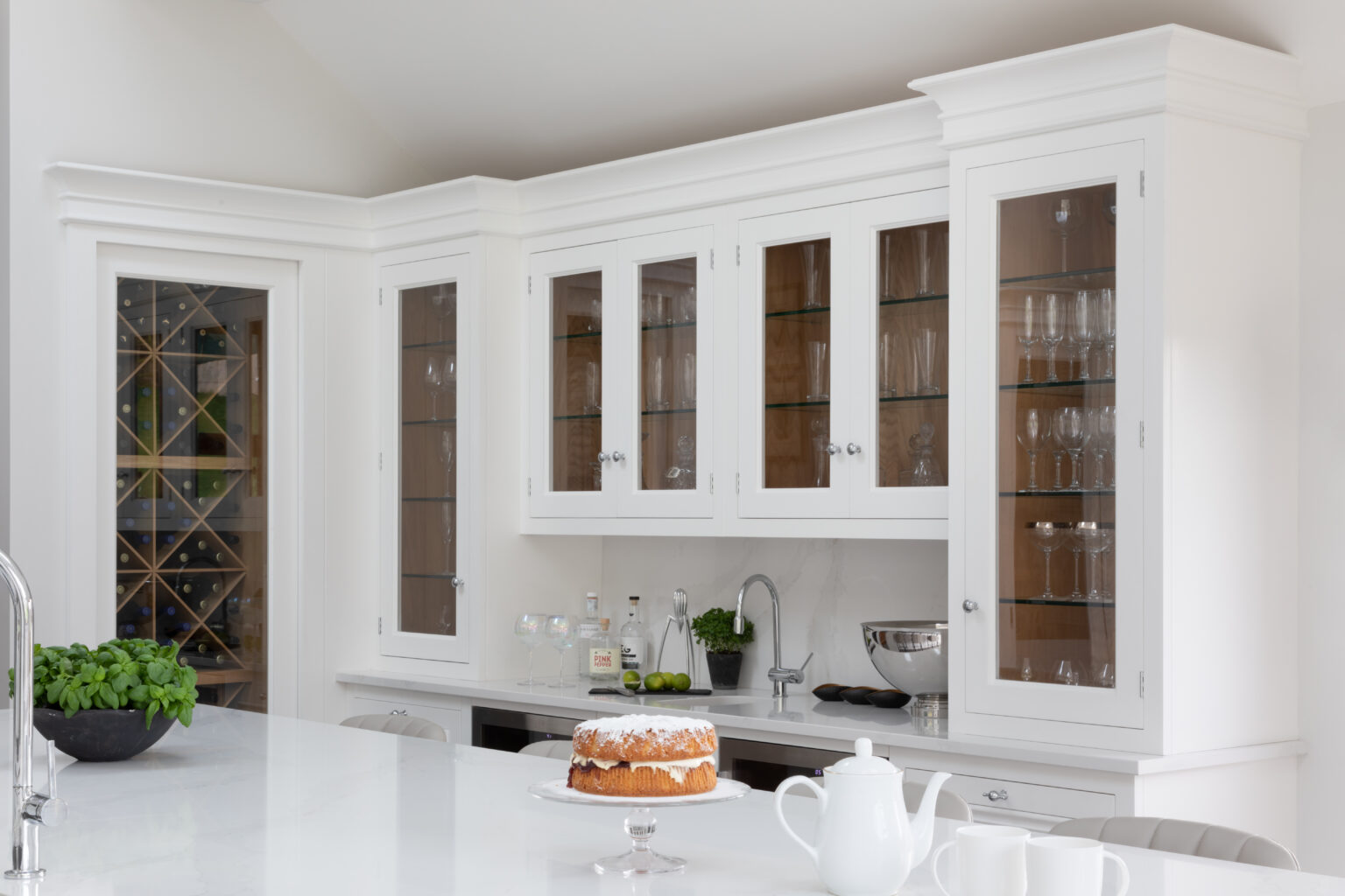 Glass-fronted white cabinets display glassware above a wet bar counter, with a diamond wine rack visible beyond