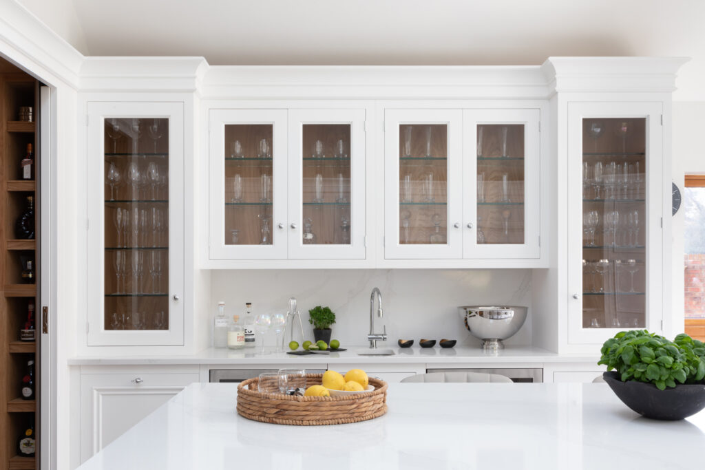 White glazed wall cabinets display glassware above a stone-effect worktop with a sink, tap and bar accessories