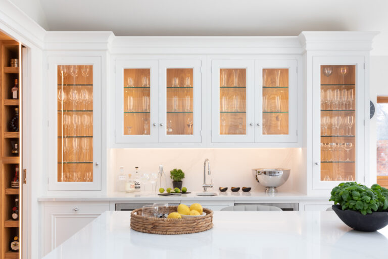 White glazed wall cabinets display glassware and decanters above a bar counter with sink, tap and bottles