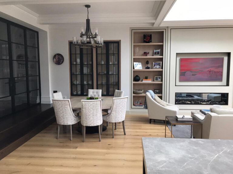 Open-plan dining and living area with round table, upholstered chairs, built-in shelving and a double-sided fireplace beyond