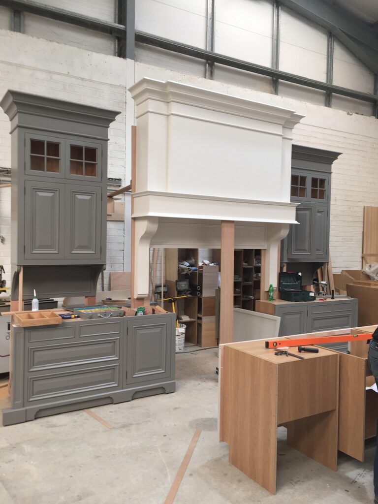 Workshop photo showing a large white panelled mantel hood between two grey painted kitchen cabinets under construction