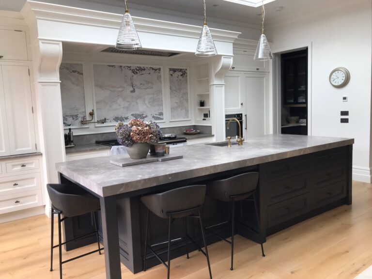 Kitchen with large dark island, stone-effect worktop, bar stools, pendant lights and a stone splashback beyond