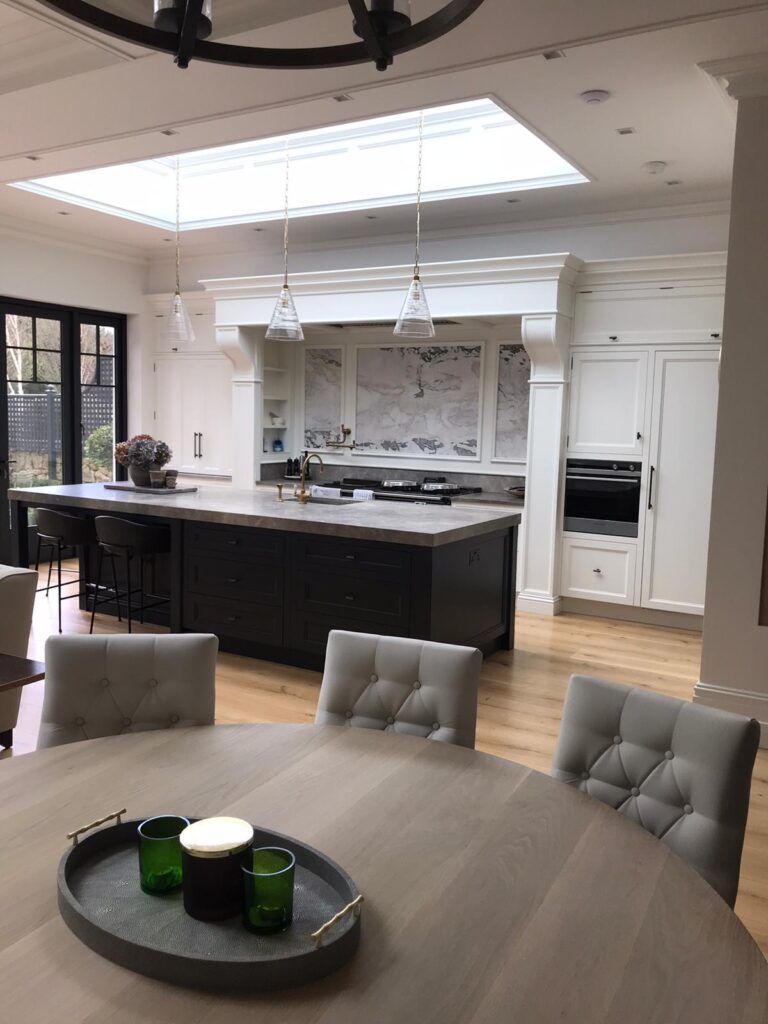 Open-plan kitchen with a dark painted island, white cabinetry, stone splashback and a round dining table in the foreground