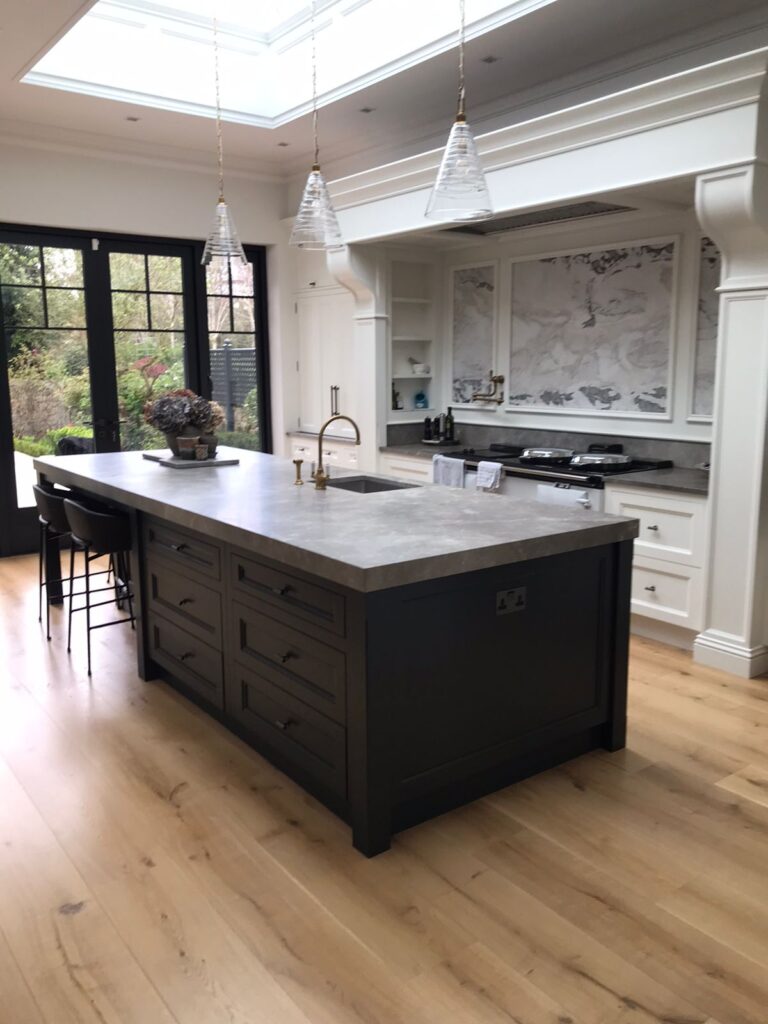 Kitchen with a dark island, stone worktop, brass tap, white cabinetry and a stone splashback beneath a skylight