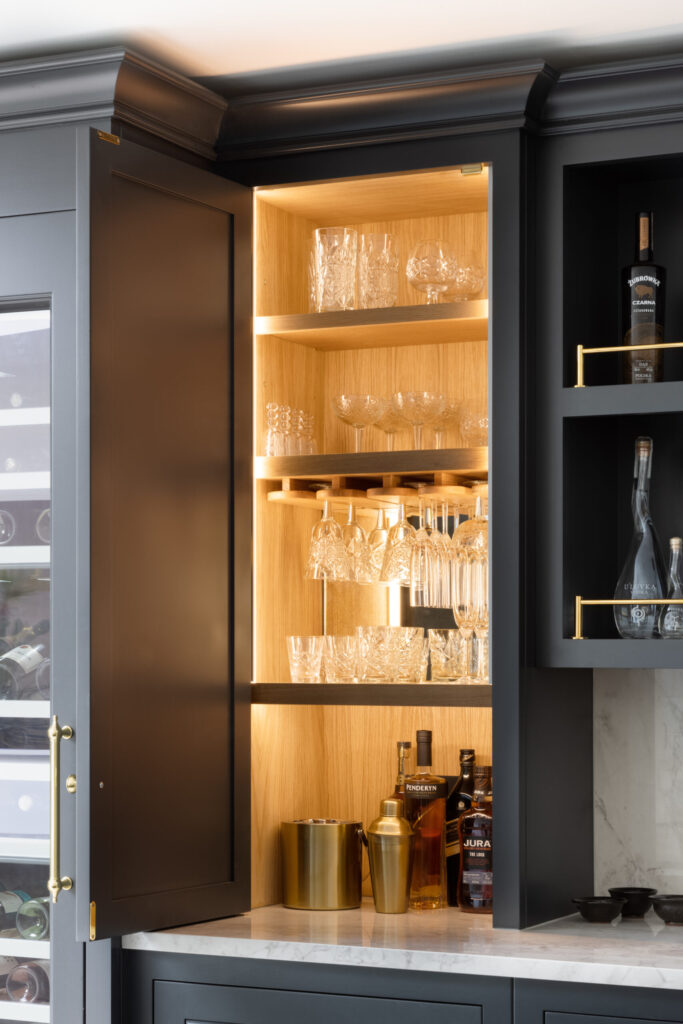 Open drinks cabinet with lit timber-lined interior, glass shelves, hanging glassware, spirit bottles and a cocktail shaker