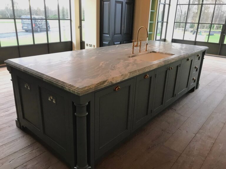 Large kitchen island with stone worktop, inset sink, brass tap and panelled cabinetry beside tall windows