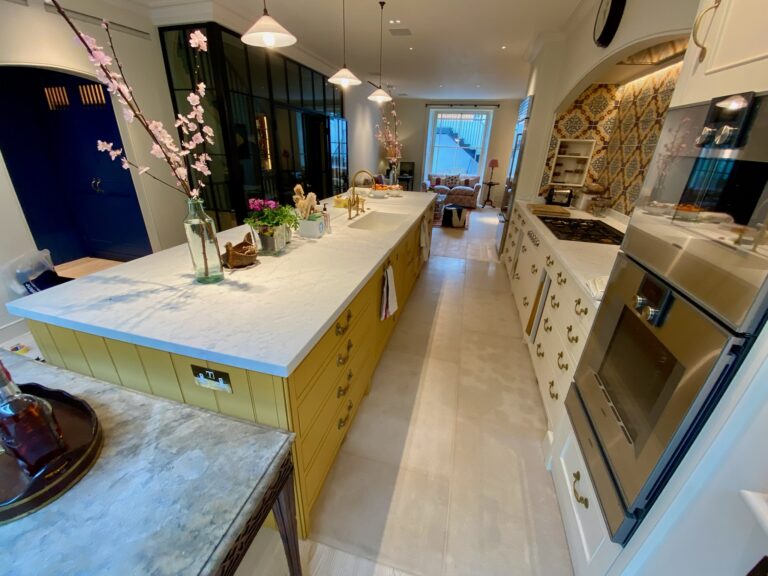 Kitchen with a long yellow island unit, stone worktop, white cabinetry and patterned tiled splashback behind the hob