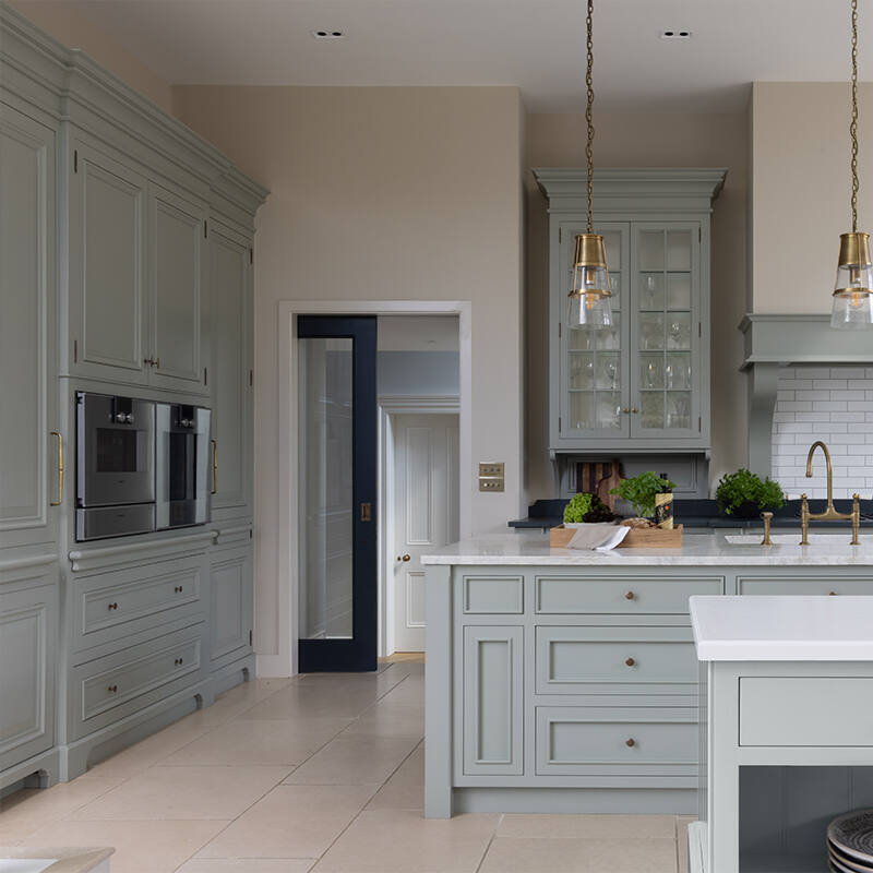 Bespoke kitchen with painted cabinetry, glass-fronted wall units, brass pendant lights and a stone-topped island