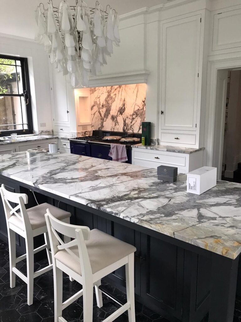 Kitchen island with veined stone worktop, dark cabinetry, white bar stools and a range cooker with matching splashback