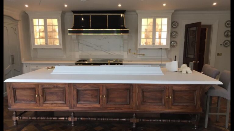 Kitchen with large wooden island unit, white worktop, black and brass range hood, and stone splashback behind the cooker